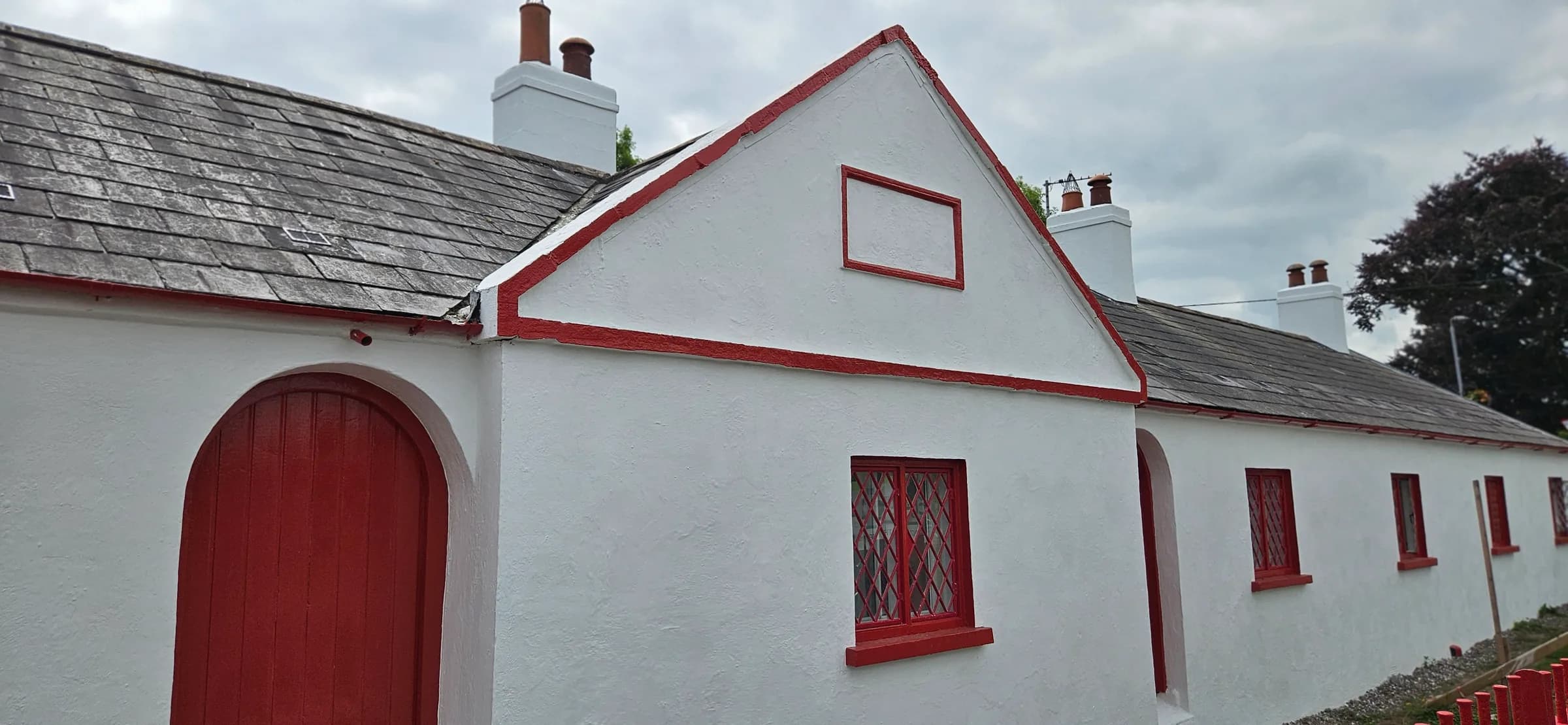 Whitewashed cottage gable with a red-painted arched timber door, red picked-out verge line and a red diamond-leaded window, under a natural slate roof