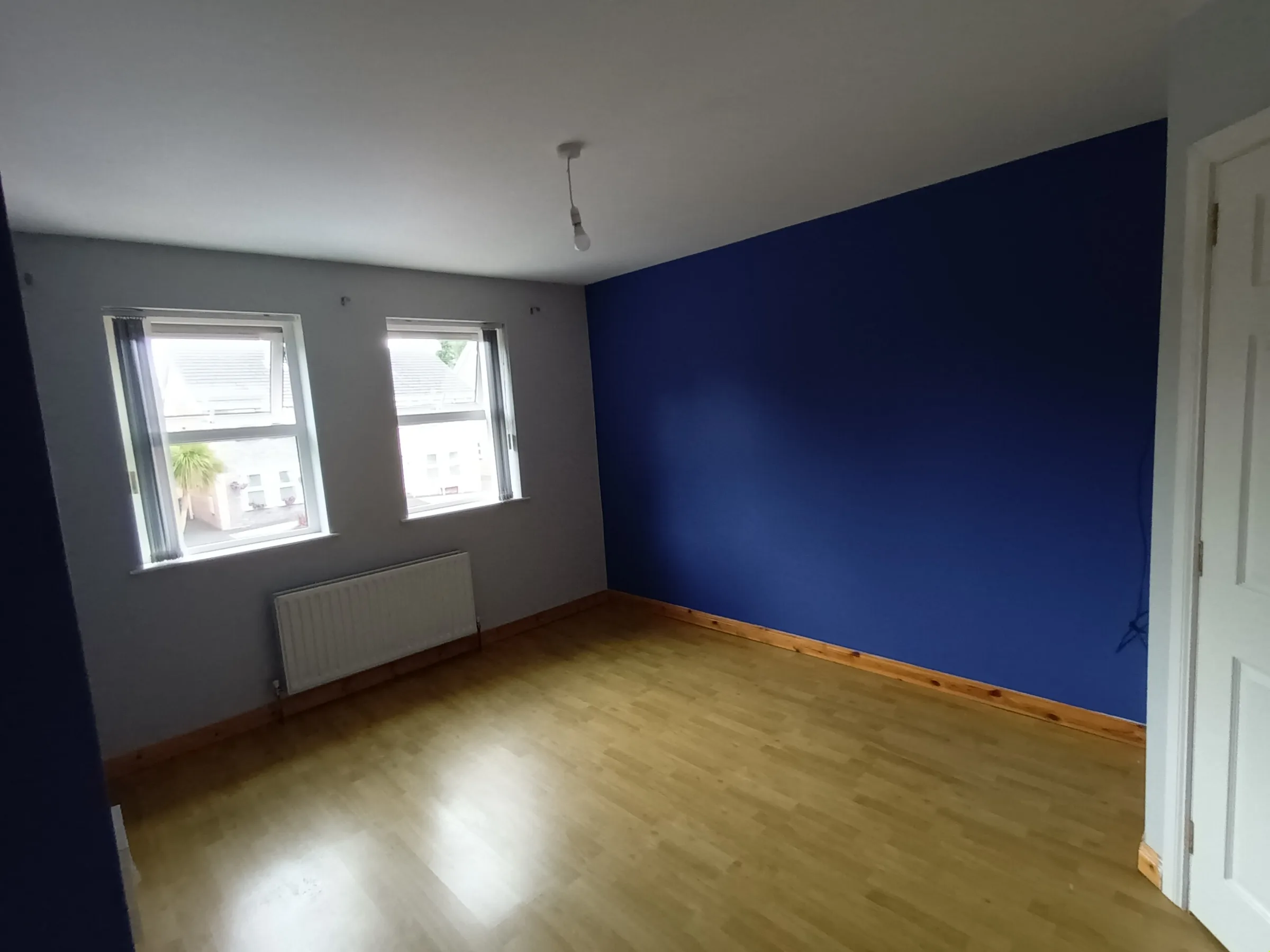 Empty bedroom with one wall in a strong cobalt blue, the remaining walls pale grey, a white ceiling, pine skirting and a laminate wood floor