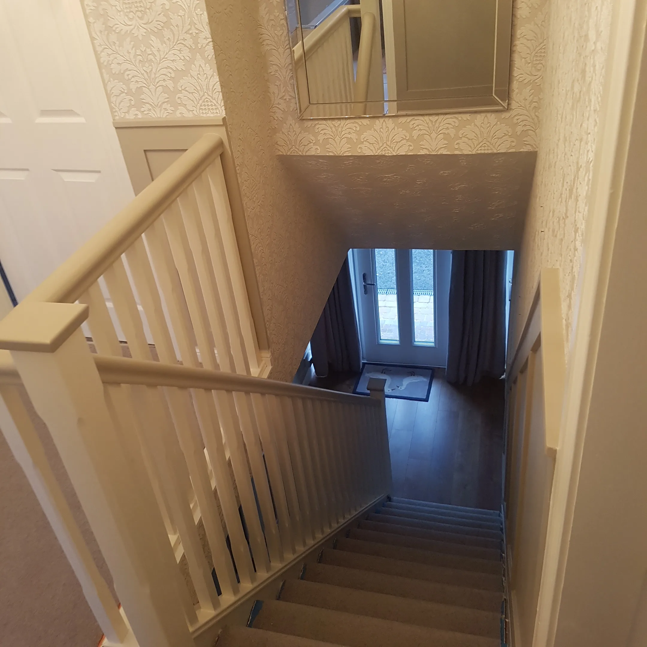 Landing and stairwell with embossed damask wallpaper painted cream, a cream spindle balustrade and handrail, looking down to a glazed front door