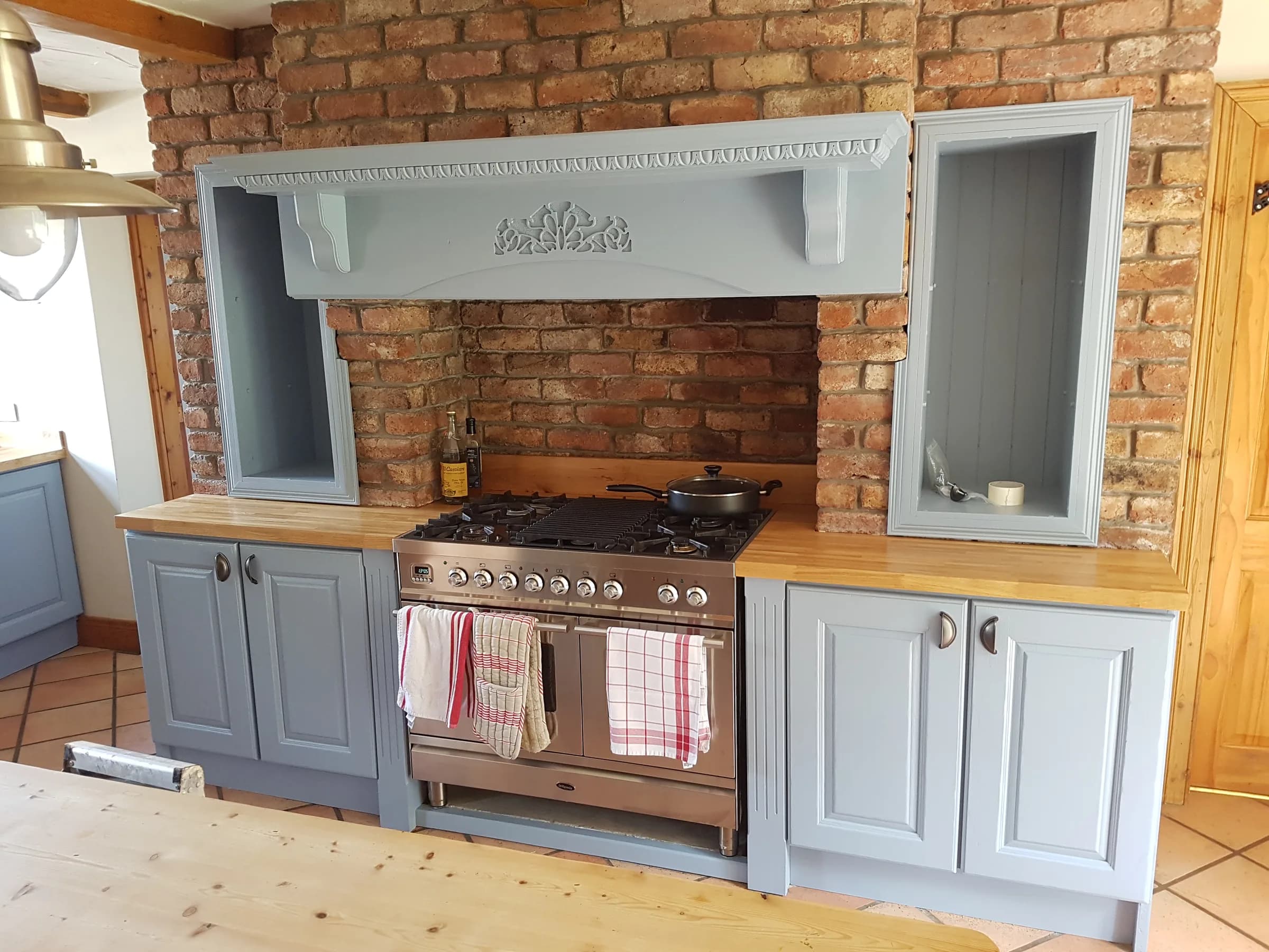 Farmhouse kitchen with an exposed red-brick chimney breast, a copper range cooker set into it, blue-grey painted raised-panel cabinets and a solid wood worktop