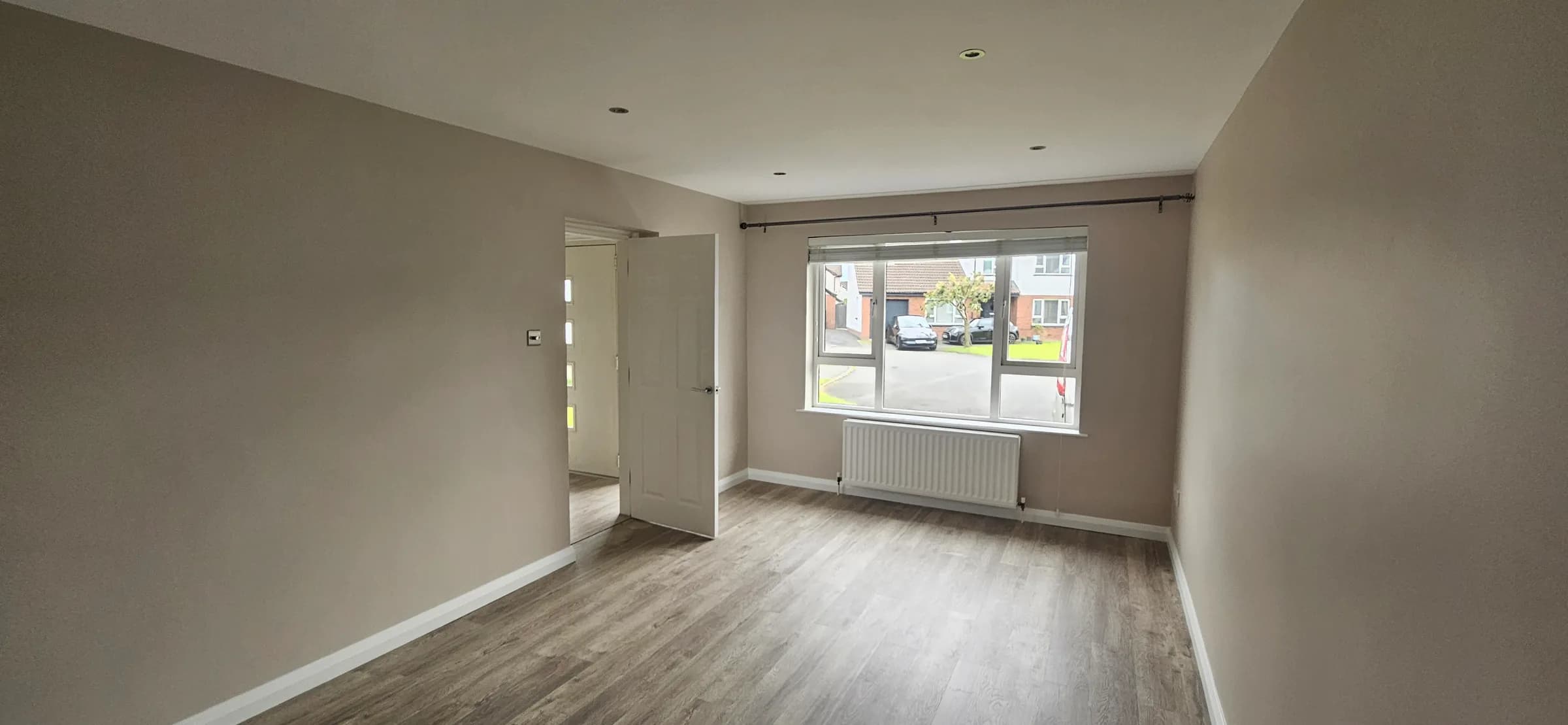 Empty room with taupe walls and white skirting, a window onto the street, a radiator beneath it and grey wood-effect flooring