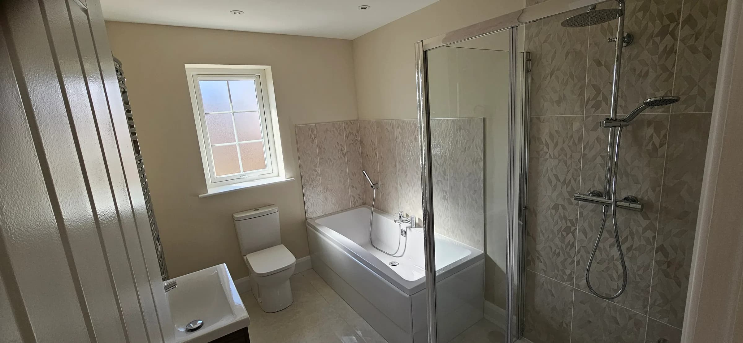 Bathroom with a bath and shower over it, patterned tiling to the shower wall, white WC and basin