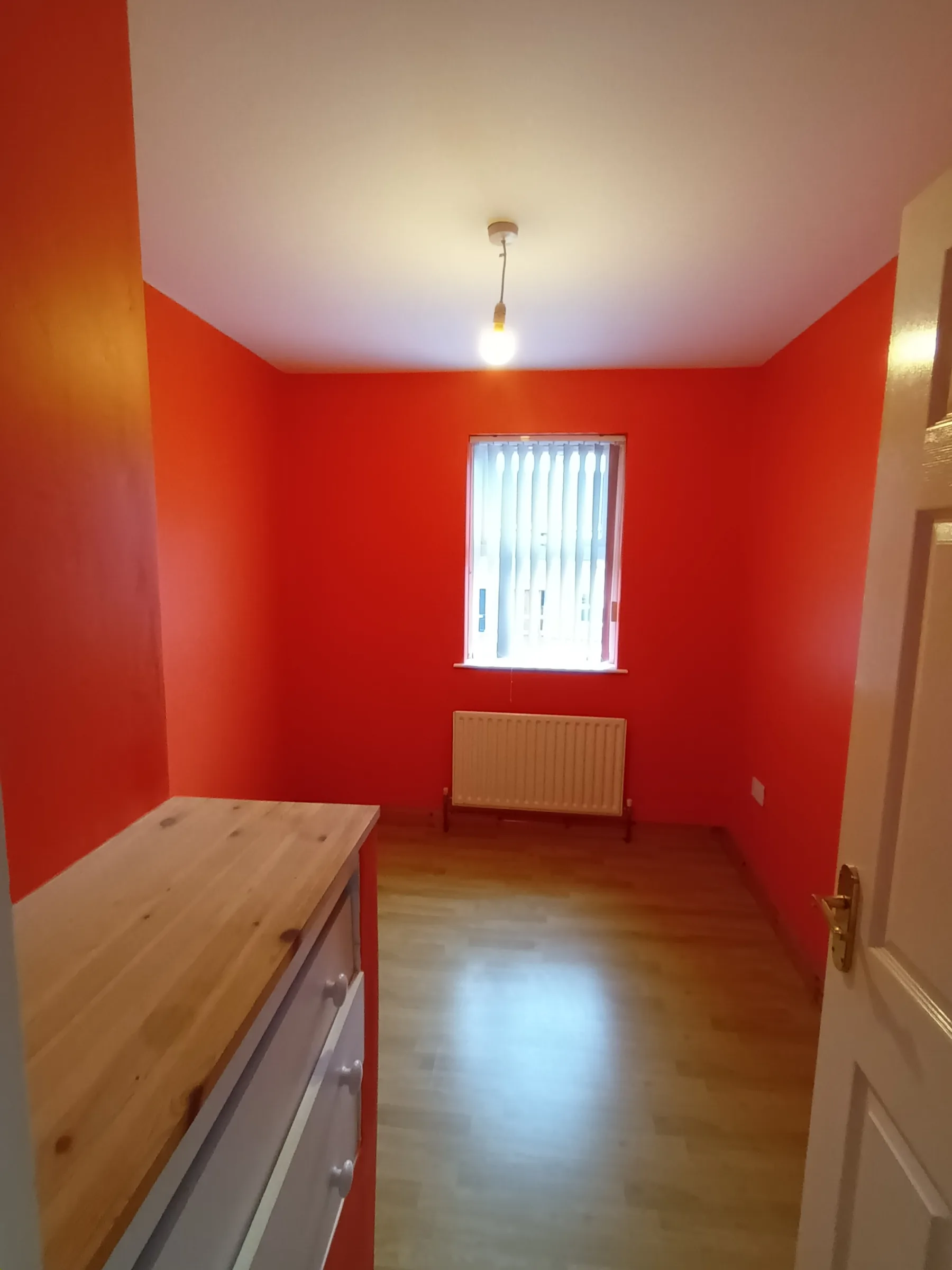 Small bedroom painted a warm orange-red on every wall, with a white ceiling, a window in white vertical blinds and a laminate wood floor