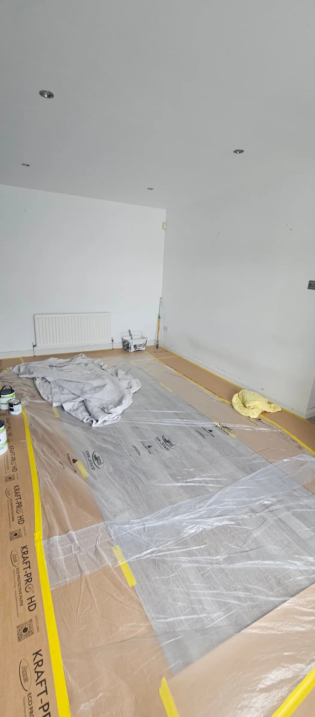 A room floor covered with Kraft-Pro paper and clear plastic sheeting, taped down along the skirting with yellow low-tack tape