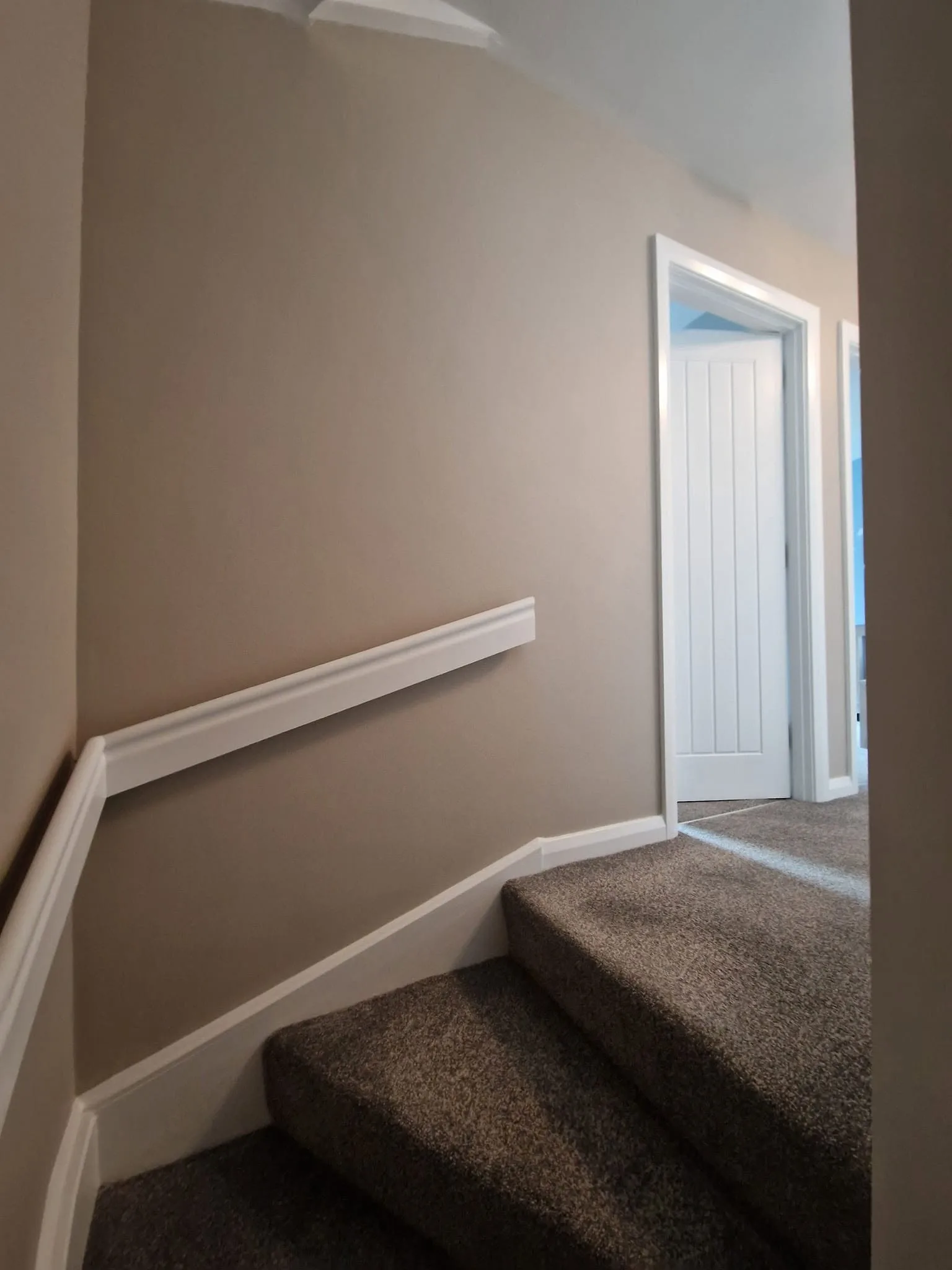 Finished upper landing in warm greige, with a white dado rail following the stair line, white skirting, a white panelled door and grey carpet