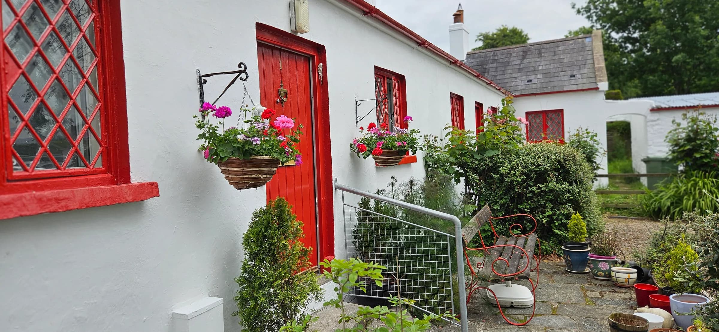 The rear of a whitewashed cottage with a red-painted door and red diamond-leaded windows, hanging baskets of geraniums either side