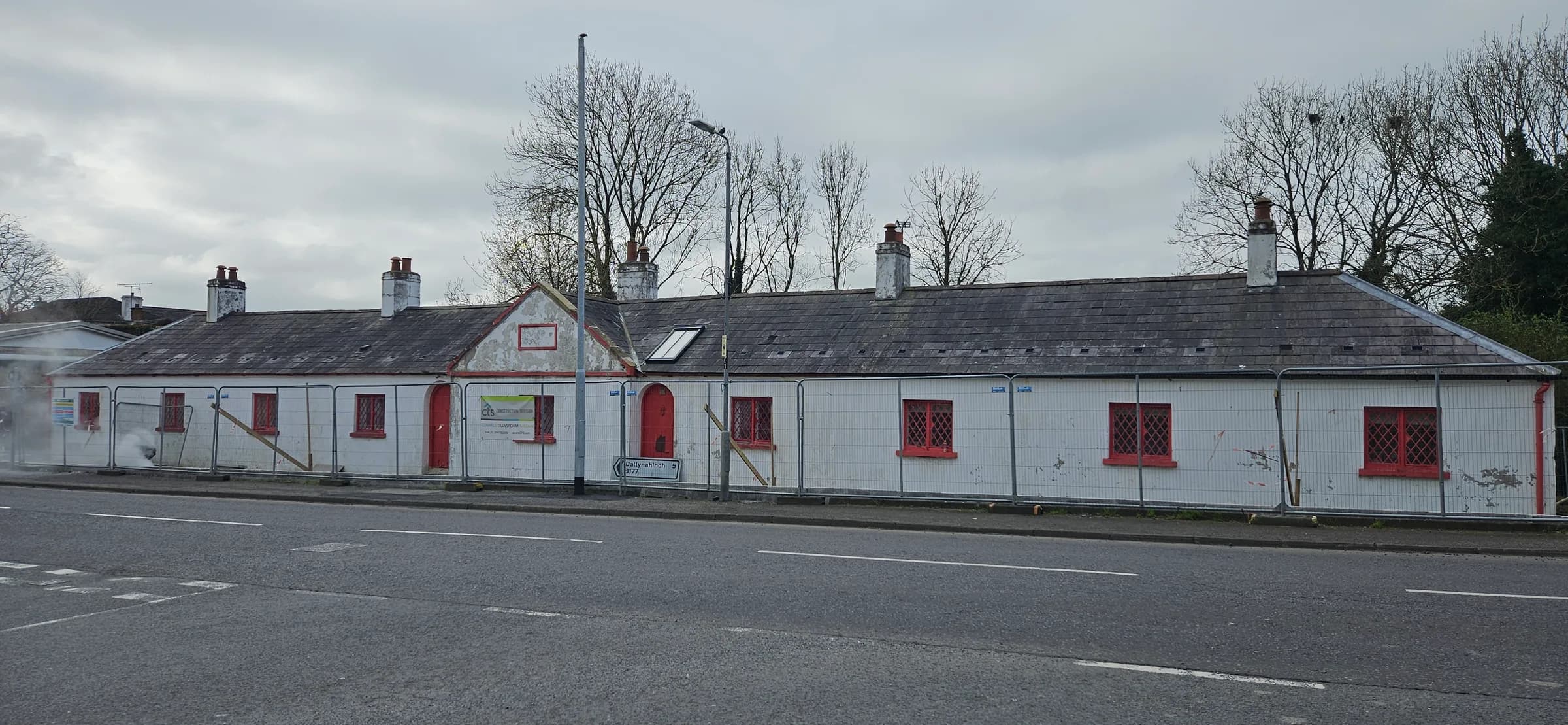 Long roadside cottage terrace behind security fencing, walls a tired grey-white, red joinery weathered