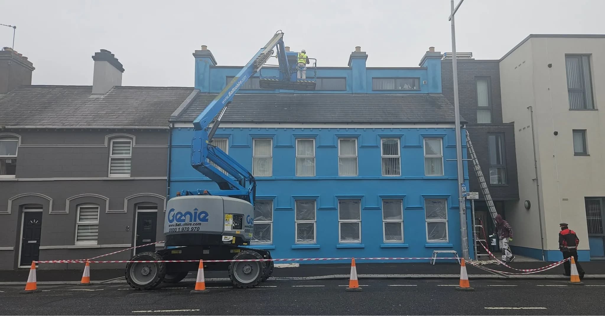 A Genie boom lift raised to roof level with an operator in the basket working on the chimney stacks, traffic cones and barrier tape closing the footpath below