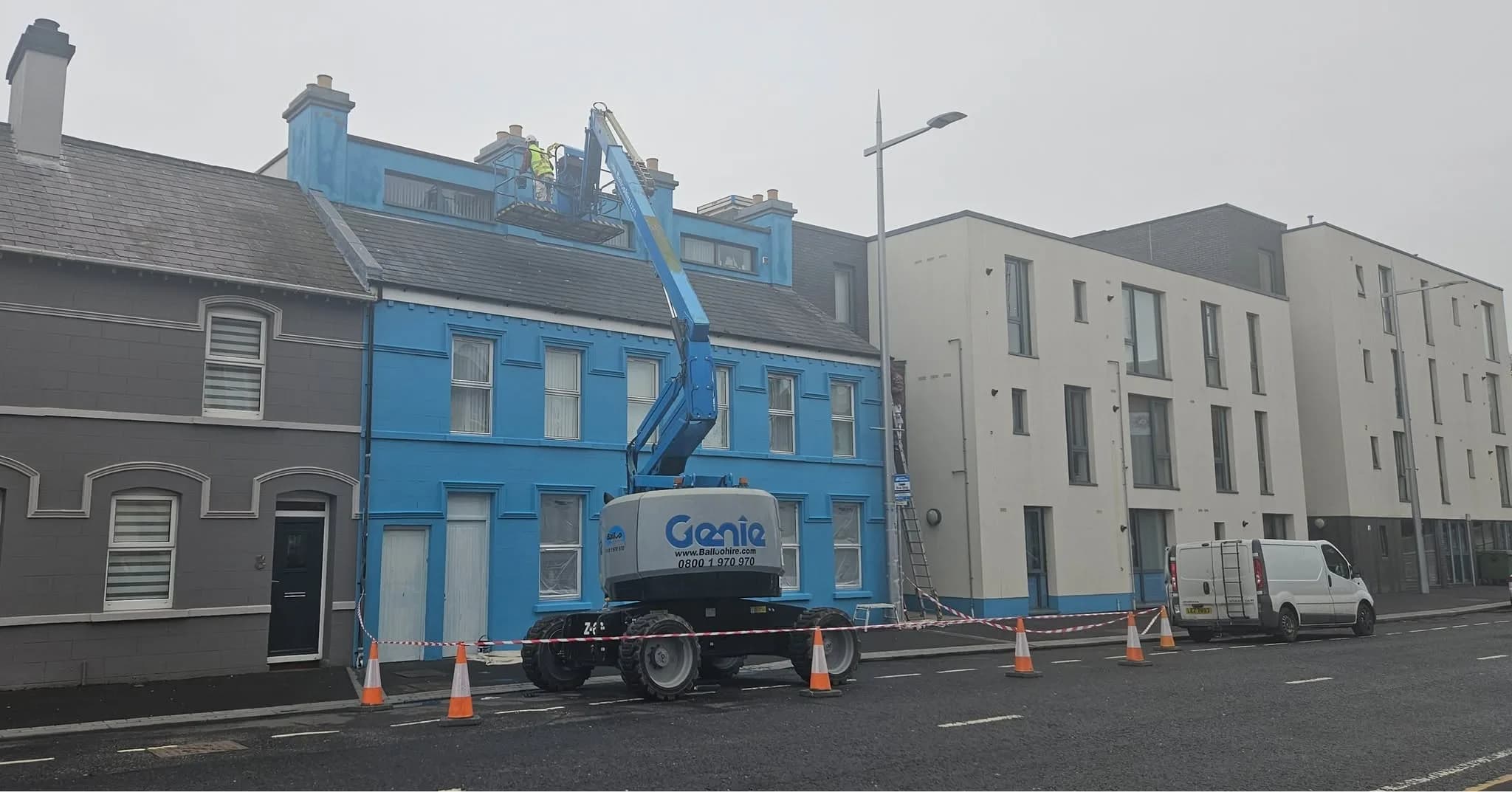 The boom lift working along the terrace from the roadway, traffic cones and barrier tape managing the footpath, a van parked beyond